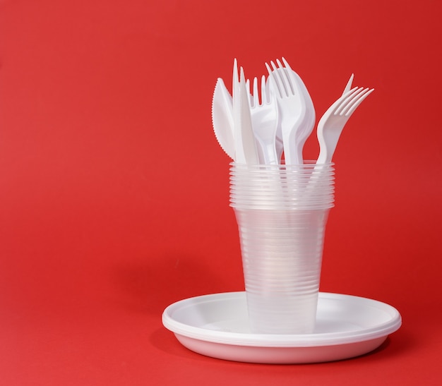 Фото Одноразовые белые пластиковые тарелки для посуды, чашки, вилки и ножи на красном фоне, набор для пикника