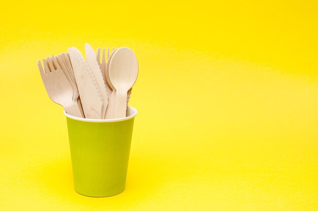 disposable wooden spoons forks and knives in a green paper cup on yellow background with copy space