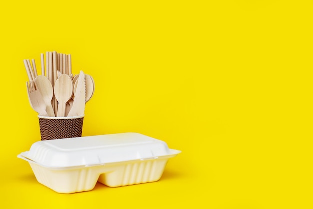 disposable wooden spoons forks and knives in a paper cup with a corn raw lunchbox container on a yellow background with copyspace recycled