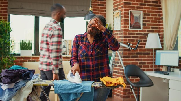 Недовольный взрослый афроамериканец занимается домашними делами без помощи партнера, генеральной уборкой. Усталая сердитая женщина гладит чистую одежду в городской квартире, работает на выходных.