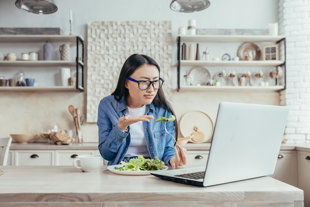 Dissatisfied asian woman with online diets woman sitting in kitchen at home eating salad for weight