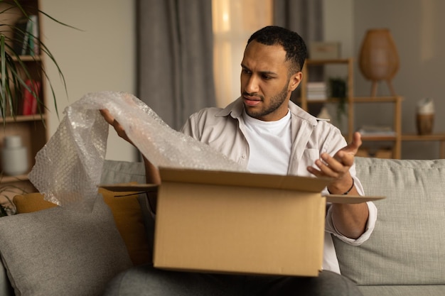 Dissatisfied black male customer opening box from online store taking out packaging unhappy with received item at home