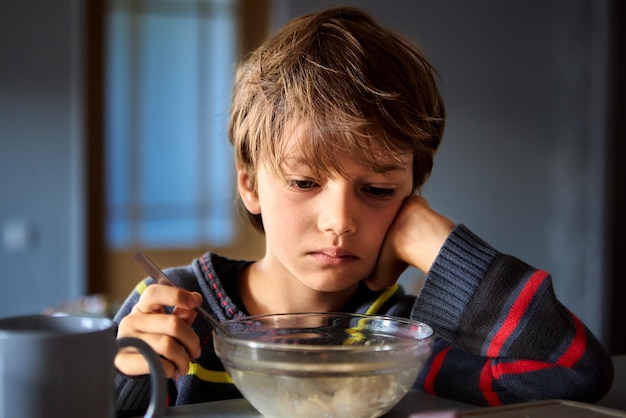 Фото Неудовлетворенный белый молодой мальчик завтракает, сидя перед прозрачной миской