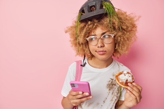 Dissatisfied curly haired young woman eats appetizing doughnut\
uses mobile phone types text messages wears protective helmet and\
white dirty t shirt isolated over pink background empty space