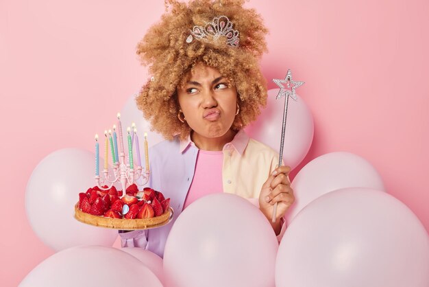 Dissatisfied curly haired young woman pouts lips looks\
unhappily aside holds festive cake and magic wand prepares for\
party and celebration poses around inflated balloons against pink\
background