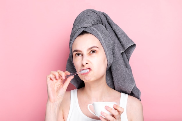 Dissatisfied and sleepy young girl drinks coffee and brushes her teeth at same time on pink space. Concept early morning rises, morning routine