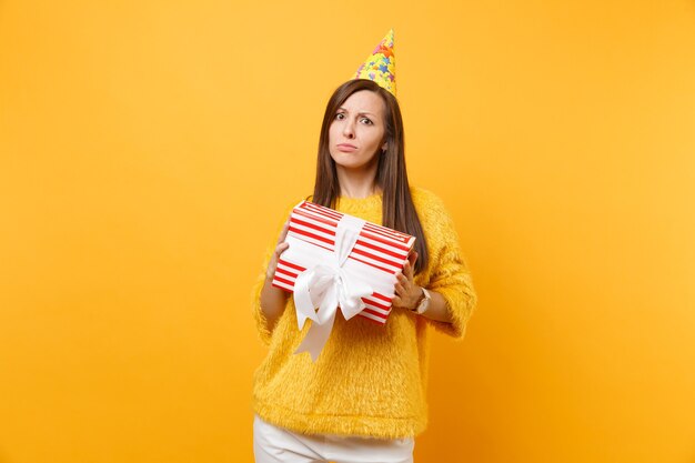 Dissatisfied upset young woman in birthday party hat holding red box with gift, present celebrating isolated on bright yellow background. People sincere emotions, lifestyle concept. Advertising area.