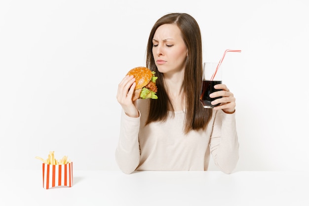写真 ハンバーガー、フライドポテト、白い背景で隔離のガラス瓶のコーラとテーブルに座っている不満の女性。適切な栄養またはアメリカの古典的なファーストフード。コピースペースのある広告エリア。
