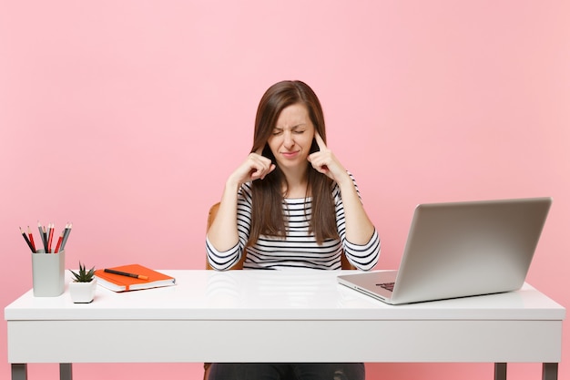 Dissatisfied woman with closed eyes does not want listen covering ears with finger sit work at white desk with contemporary pc laptop