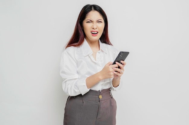 A dissatisfied young Asian woman looks disgruntled wearing white shirt irritated face expressions holding her phone