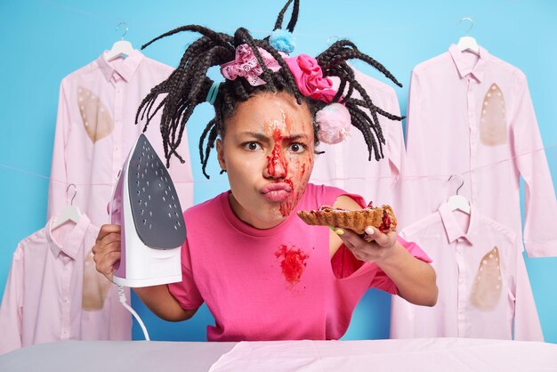 Dissatisfied young housewife with braided hair eats delicious\
sweet homemade pie does ironing at home dressed casually poses\
against ironed shirts on hangers blue background household\
concept