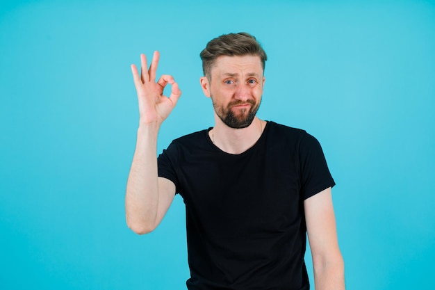 Dissatisfied young man is showing okay gesture by raising up his hand on blue background