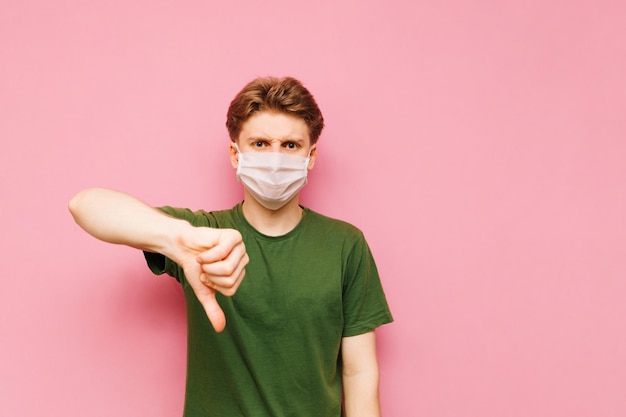 Dissatisfied young man stands on a pink background in a medical mask and shows thumbs up Guy in a gauze mask on his face shows a dislike gesture and looks at the camera with a sad face