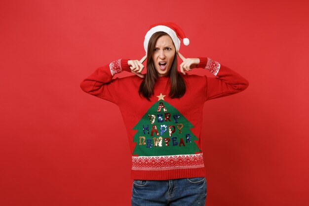 Dissatisfied young Santa girl in knitted sweater, Christmas hat covering ears with fingers isolated on bright red background. Happy New Year 2019 celebration holiday party concept. Mock up copy space.