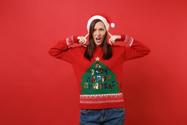 Foto ragazza insoddisfatta di santa in maglione lavorato a maglia, cappello di natale che copre le orecchie con le dita isolate su sfondo rosso brillante. felice anno nuovo 2019 celebrazione festa concetto. mock up copia spazio.