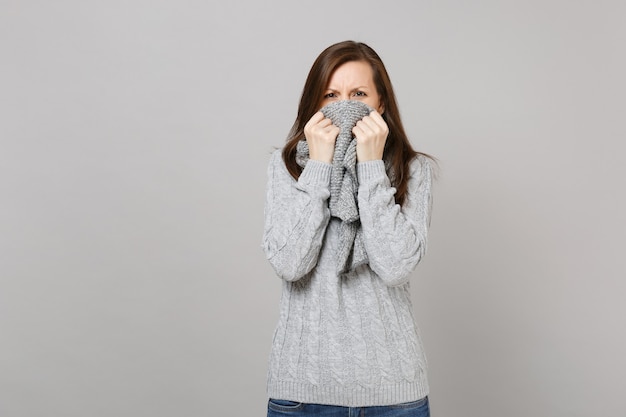 Dissatisfied young woman in gray sweater covering mouth with scarf isolated on grey background in studio. Healthy fashion lifestyle people sincere emotions, cold season concept. Mock up copy space.