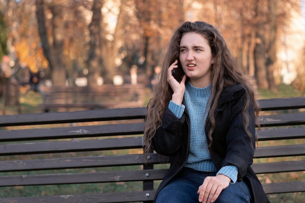 Dissatisfied young woman sits in park on bench and talks on phone and gesticulating Girl in autumn park