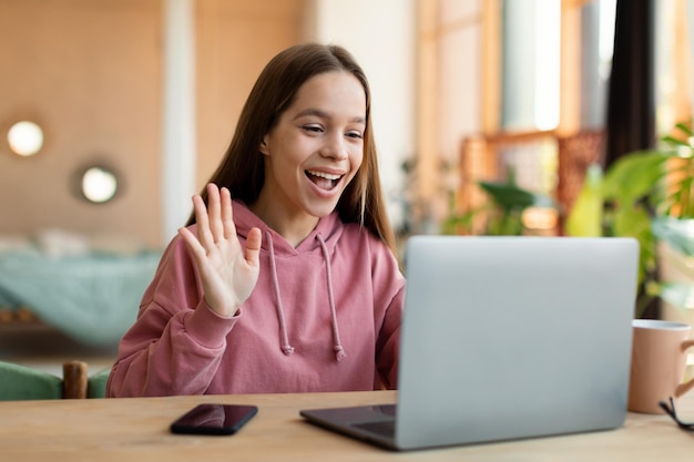 Concetto di comunicazione a distanza ritratto di una ragazza adolescente allegra che parla con il suo tutor o con gli amici che salutano la webcam del laptop