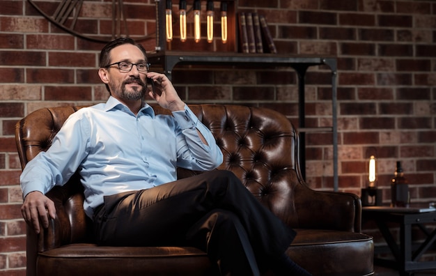 Distance communication. Handsome delighted good looking businessman putting the smartphone to his ear and listening to his interlocutor while being in his study room