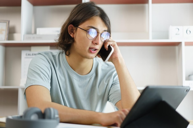 Distance education concept Teen boy talking on phone and using tablet to lesson class online