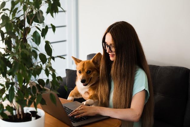 원격 교육 안경을 쓴 여학생이 집에서 사랑스러운 Corgi 강아지와 함께 온라인으로 공부하고 있습니다.