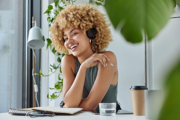 Distance education and studying online concept positive curly\
haired woman listens audio lecture via headphones connected to\
digital tablet writes down notes with pen drinks takeaway\
coffee