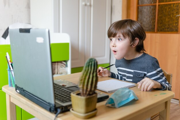 코로나 바이러스가 유행하는 동안 어린이를위한 원격 교육. 한 소년이 테이블에 앉아 인터넷에서 교사의 작업을 수행합니다.