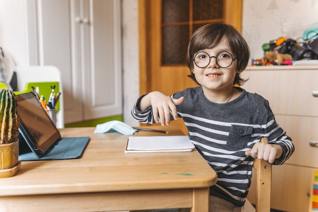 Дистанционное обучение детей во время эпидемии коронавируса. Мальчик сидит за столом и выполняет задания учителя в Интернете.