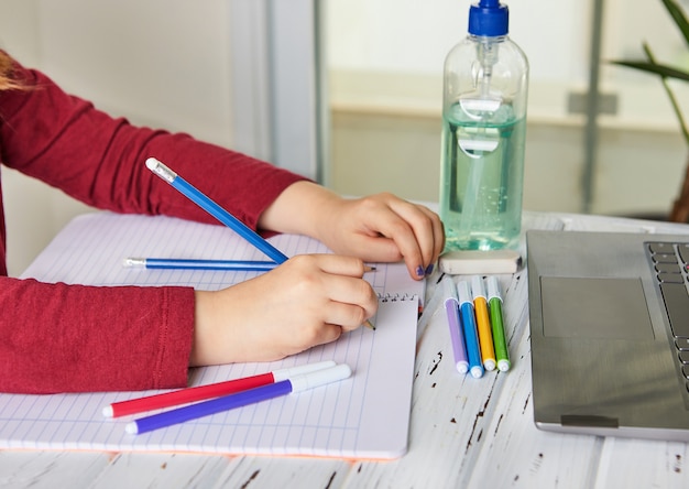 Distance learning online education. Schoolgirl studying at home with a laptop and doing school homework. Training books and colored felt-tip pens on the table, gel with alcohol 70 percent