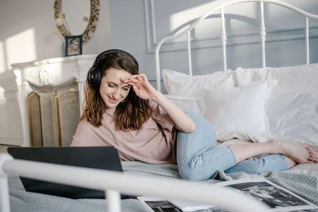 Distance learning online education and work. Happy woman in casual with the headphones working on a laptop remotely from home on bed.