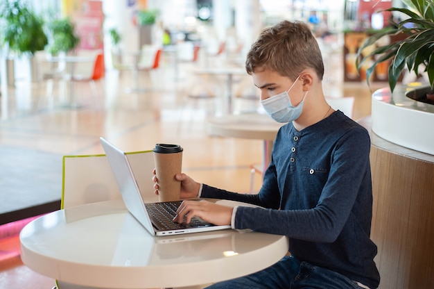 Дистанционное обучение. Мальчик-подросток в медицинской маске сидит за столиком в кафе с ноутбуком и кружкой горячего напитка. Фото высокого качества