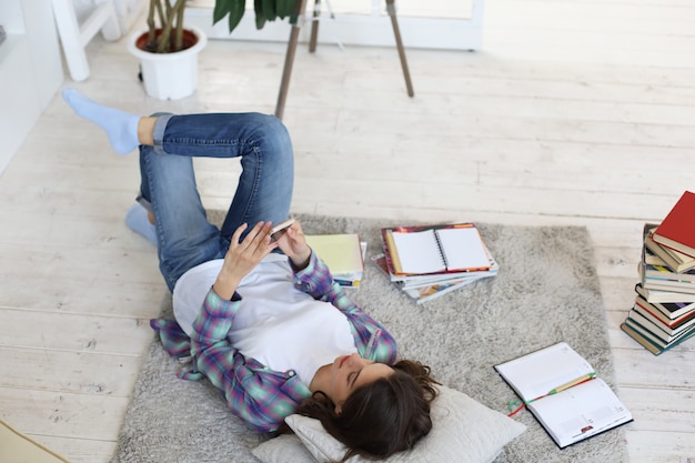 Insegnamento a distanza. giovane studentessa che studia a casa, si prepara per gli esami universitari, sdraiata sul pavimento contro l'accogliente interno domestico, circondata da una pila di libri