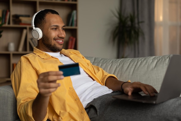 Distance shopping concept Cheery young black guy with laptop and credit card making online order from home