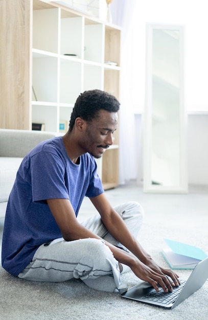 Distance work Casual man Online connection Smiling guy in home look typing laptop sitting floor in light room interior