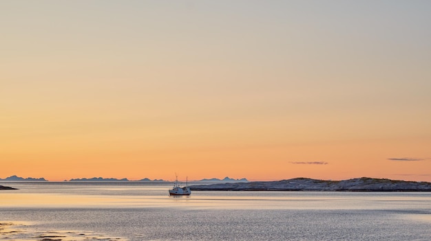 ノルウェーの夏の日の夜明けの間に、日没時に水中の山の近くの海にある遠くの帆船海のビーチまたは遠くにある丘の近くに船舶を持つ大きな湖