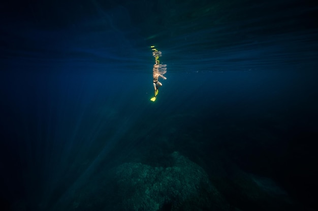 Distant unrecognizable male diver in yellow flippers swimming under dark blue water of deep sea during holidays