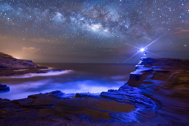 Photo distant view of man holding illuminated flashlight on rock at shore against star field