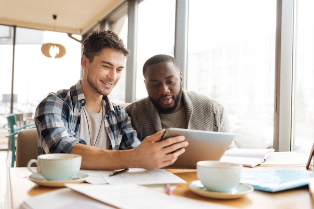 写真 気が散る注意。若い魅力的な男は、カフェに座っている間、彼の新しいタブレットで彼の仲間にいくつかのコンテンツを見せています。