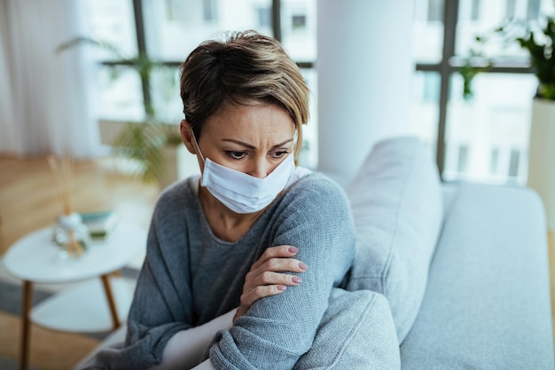 Distraught woman sitting on the sofa while wearing protective face mask and thinking during COVI19 epidemic