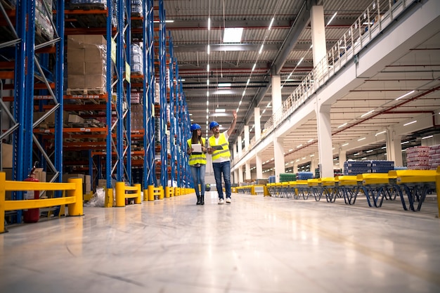 Distribution warehouse interior with workers wearing hardhats and reflective jackets walking in storage area.