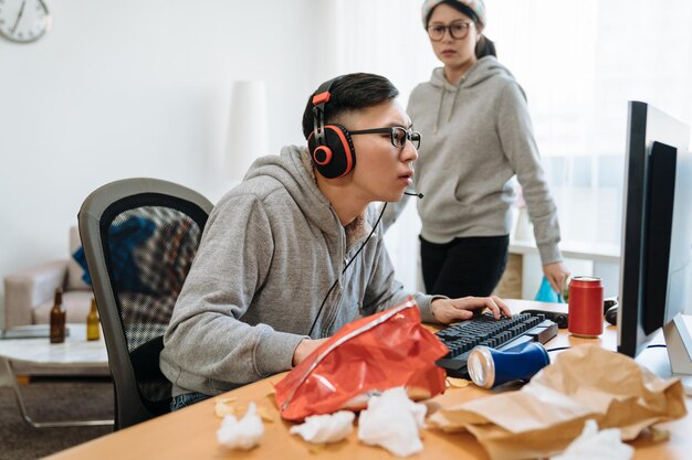 distrustful girlfriend annoyed with boyfriend technology addiction. bokeh angry young asian woman staring at guy and walking clean up while man playing on computer online game in messy room at home