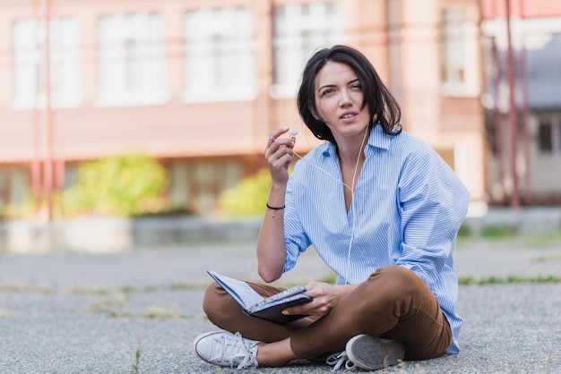 Disturbed European beautiful woman while sitting at sidewalk studying and making notice on notebook  planner Young woman with earphone listen and preparing for exam