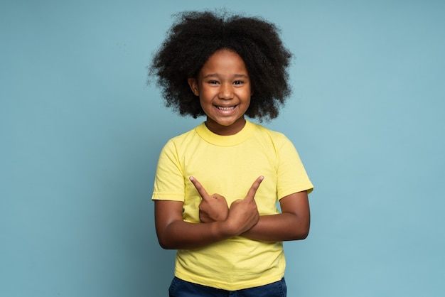 Foto dit ben ik. portret van een gelukkig voorschools krullend meisje in t-shirt dat vreugdevol naar de camera kijkt en naar zichzelf wijst, trots op eigen succes. indoor studio-opname geïsoleerd op blauwe achtergrond