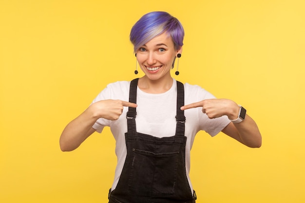 Dit ben ik. Portret van een vrolijke hipstervrouw met violet kort haar in een denimoverall die zichzelf wijst, glimlacht en trots uitdrukt op persoonlijke prestaties. geïsoleerd op gele achtergrond studio shot