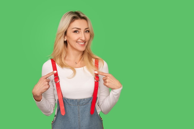 Dit ben ik portret van zelfverzekerde ambitieuze volwassen vrouw in denim overalls wijzend naar zichzelf en glimlachend kijken trots op prestatie succes indoor studio shot geïsoleerd op groene achtergrond