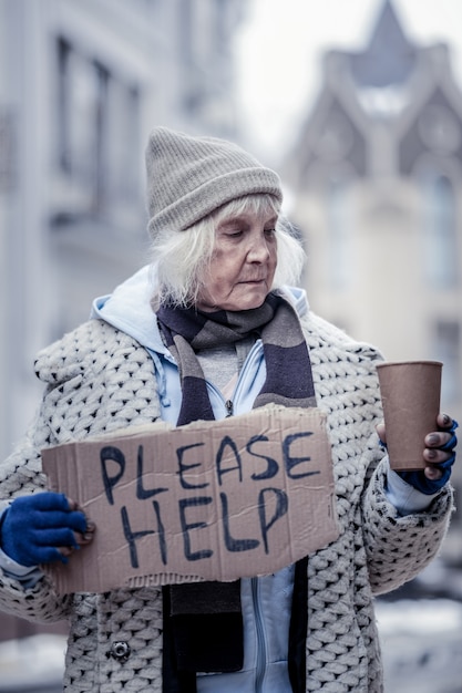 Dit is armoede. Ongelukkige ongezellige vrouw die op straat staat terwijl ze in de beker met munten kijkt