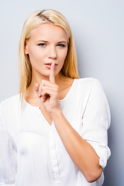 Dit is gewoon tussen jou en mij! close-up van een aantrekkelijke jonge vrouw die naar de camera kijkt en de vinger op de lippen houdt terwijl ze tegen een grijze achtergrond staat