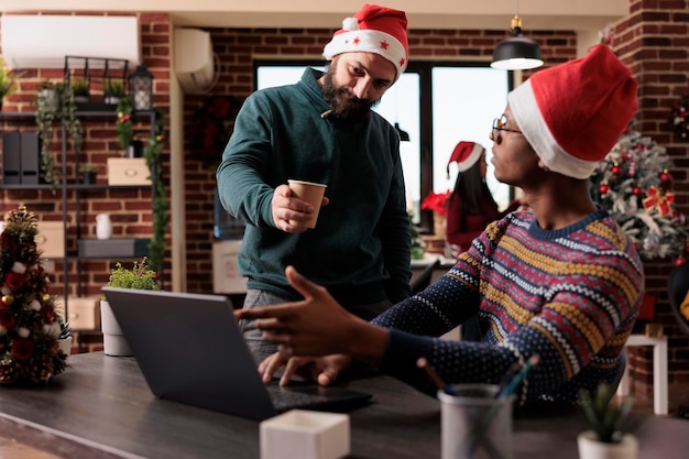 Divers team van zakenlieden in feestelijke ruimte die op laptop werken om rapport te plannen, met behulp van gegevens in kantoor versierd met feestelijke ornamenten en kerstboomverlichting. Werken tijdens het winterseizoen.