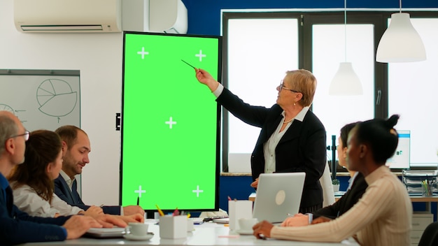 재무 통계를 분석하는 회의 테이블에 앉아 있는 다양한 비즈니스 팀은 수석 리더가 설명하는 동안 녹색 화면 디스플레이를 보고 있습니다. 크로마 키 데스크탑에서 프로젝트를 계획하는 다민족 근로자