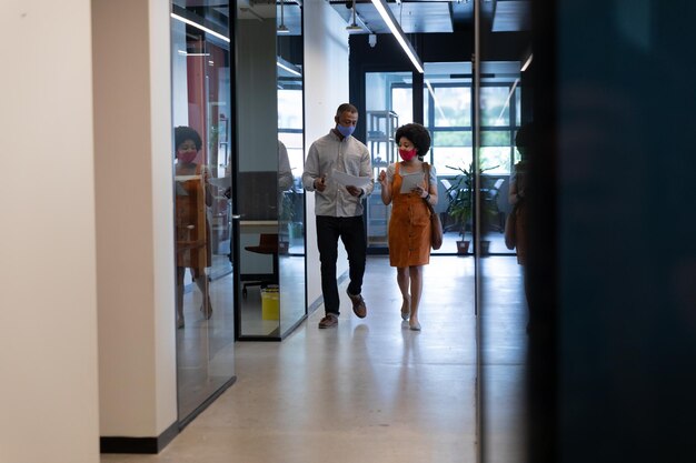 Diverse businessman and businesswoman wearing face masks checking documents in office. social distancing in business office workplace during covid 19 coronavirus pandemic.